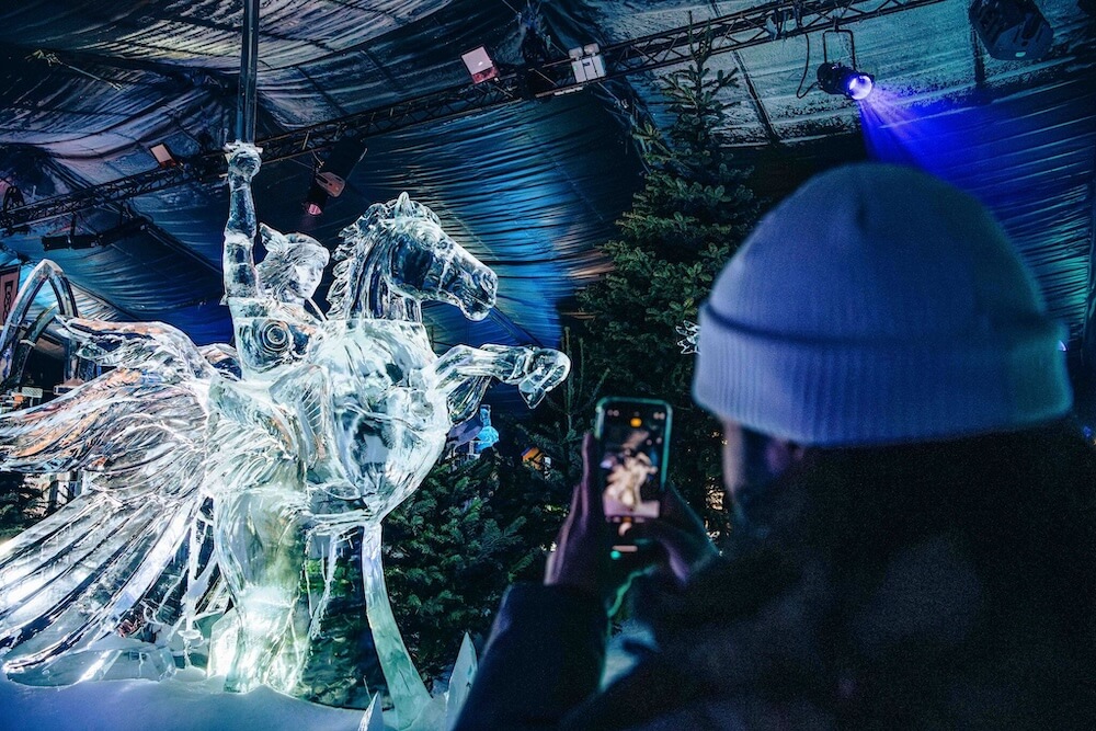 A person in a white beanie photographs a detailed ice sculpture of a winged horse with a rider, set against a backdrop of trees and blue lighting.