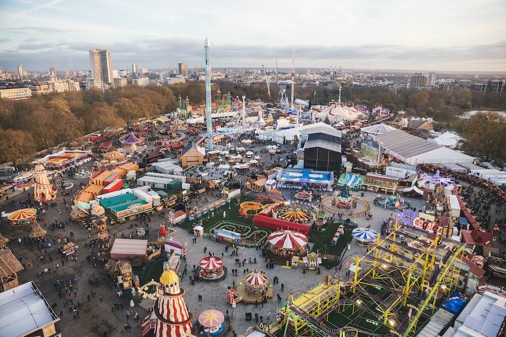 Aerial view of a bustling winter fairground with vibrant rides, colourful tents, and crowds of people. Trees and city skyline in the background. Festive atmosphere.