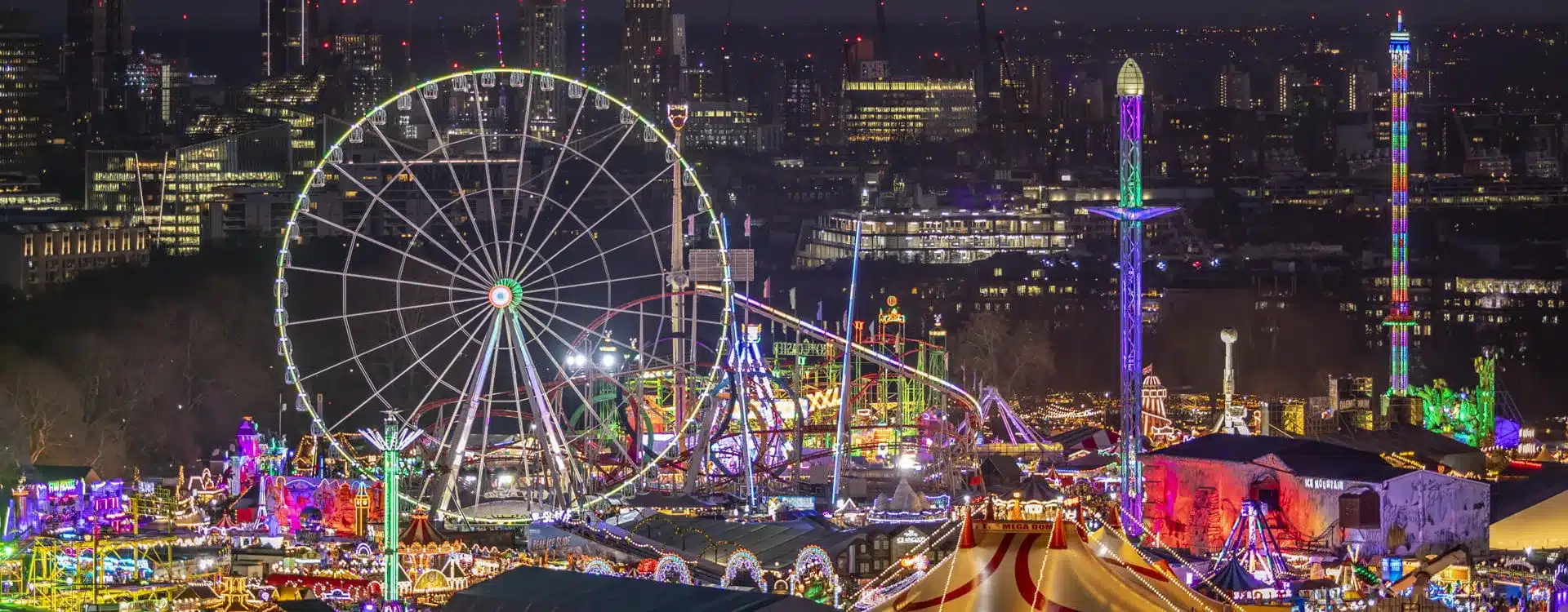 Illuminated fairground at night with a lit Ferris wheel, vibrant stalls, and colorful rides surrounded by a bustling cityscape in the background.