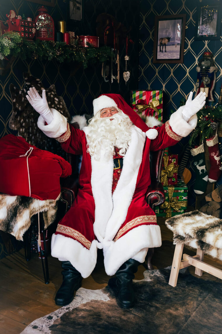 Santa Claus is sitting in a cozy, festive room with arms wide open, surrounded by Christmas decorations and gifts. The atmosphere is joyful and welcoming.