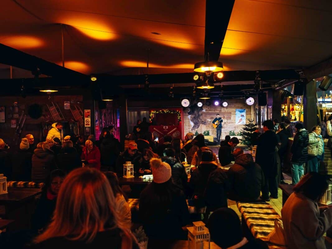 A cozy winter scene at the Explorers Rest with warm lighting shows a musician playing guitar on stage. A crowd of people, bundled in coats and hats, enjoys the lively atmosphere.