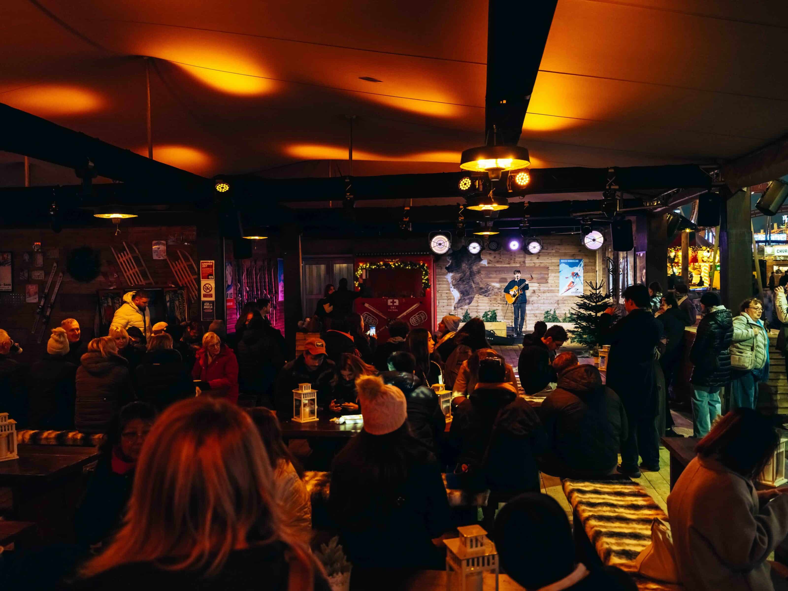 A cozy winter scene at the Explorers Rest with warm lighting shows a musician playing guitar on stage. A crowd of people, bundled in coats and hats, enjoys the lively atmosphere.