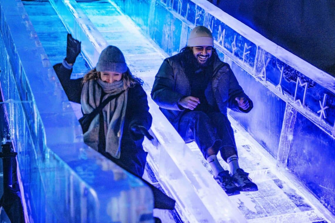 Two people dressed warmly in coats and hats joyfully slide down the glowing blue real ice slide, conveying excitement and winter fun in a festive atmosphere.
