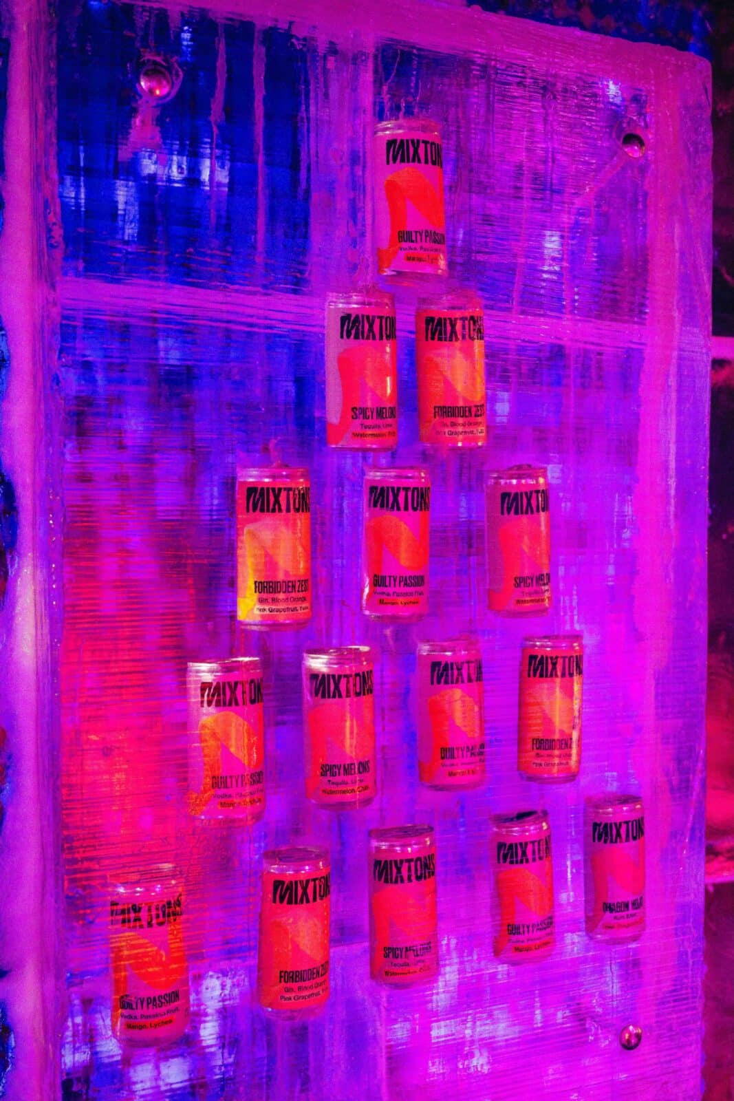 Cans of "Mixtons Cocktails" drinks are arranged in a pyramid shape on a clear shelf, illuminated by vivid pink and purple lights, creating a vibrant, energetic display.