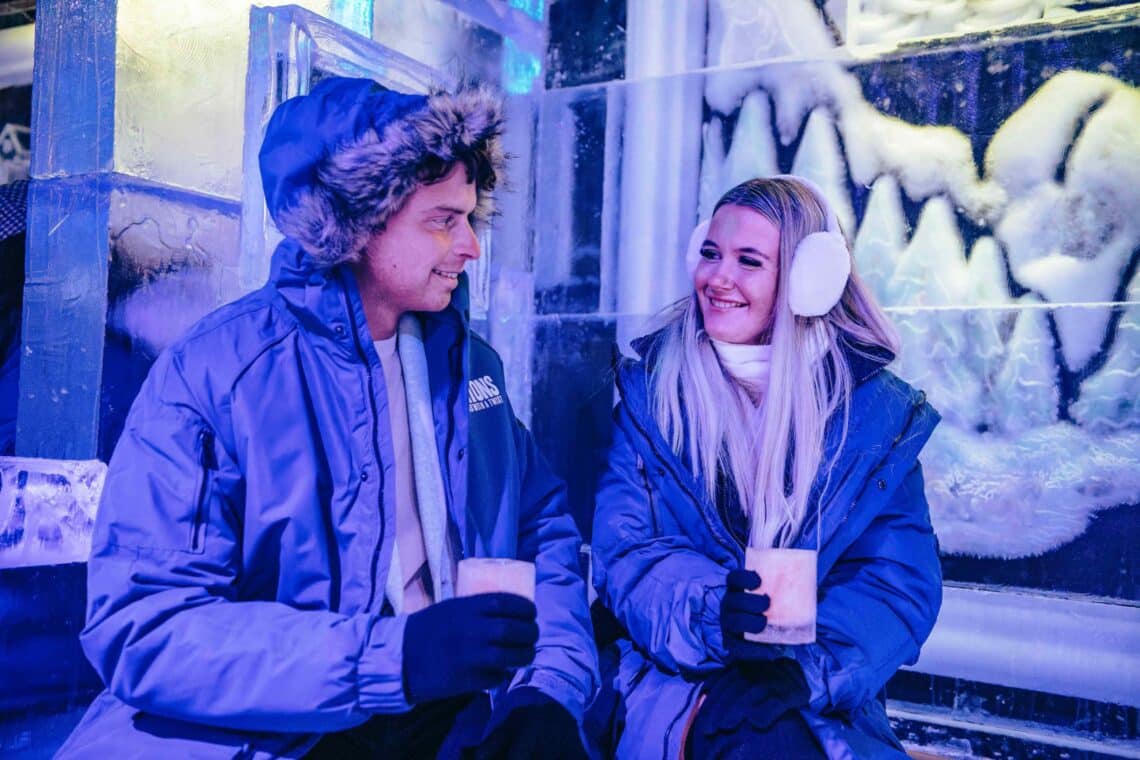 Inside Bar Ice, a man and woman in winter coats and earmuffs sit on ice seats, holding drinks. They smile at each other in a warmly lit ice bar, conveying a cozy atmosphere.
