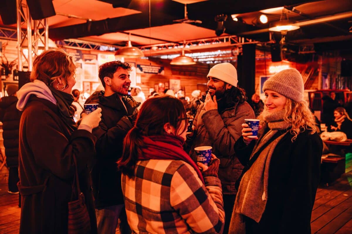 A group of five people in winter clothing, holding festive cups, engage in cheerful conversation indoors. Warm lighting and cozy décor create a festive atmosphere.