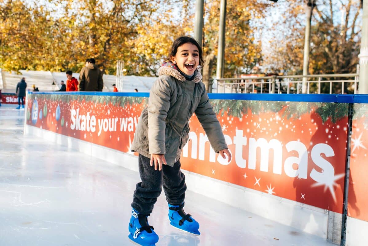 A joyful child wearing a winter coat and blue ice skates glides on an ice rink. Behind, a festive "Skate your way to Christmas" sign and autumn trees.