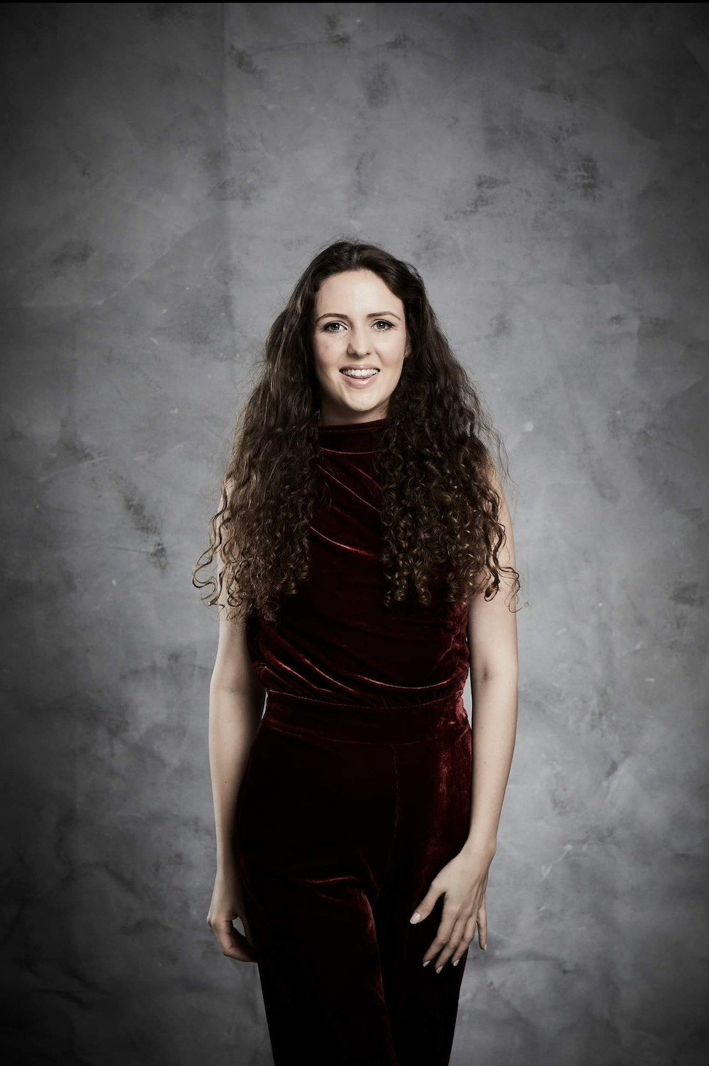 Mariska Gandey Briggs, smiling with long curly hair, wears a dark red velvet jumpsuit against a textured grey studio backdrop.