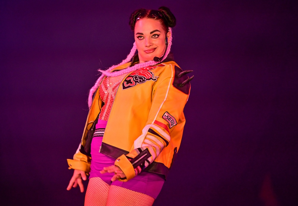 La Loka performs on stage wearing a yellow and pink jacket with 'Seoul' patch, pink space buns, and a headset microphone, posing against a dark purple lit background.