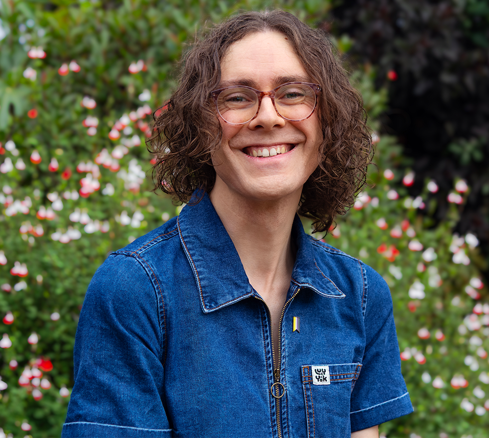 Harry Woodgate, a smiling person with curly brown hair and glasses, wearing a blue denim shirt with a pin badge, stands in front of flowering shrubs.