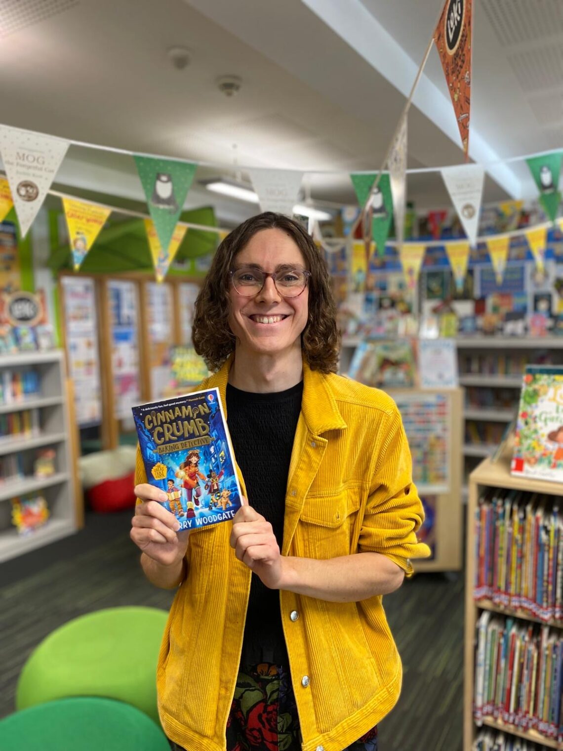A smiling bookstore employee in a bright yellow jacket holds up “Cinnamon Crumb” amid colourful shelves and festive pennants, creating a warm, cheerful feel.