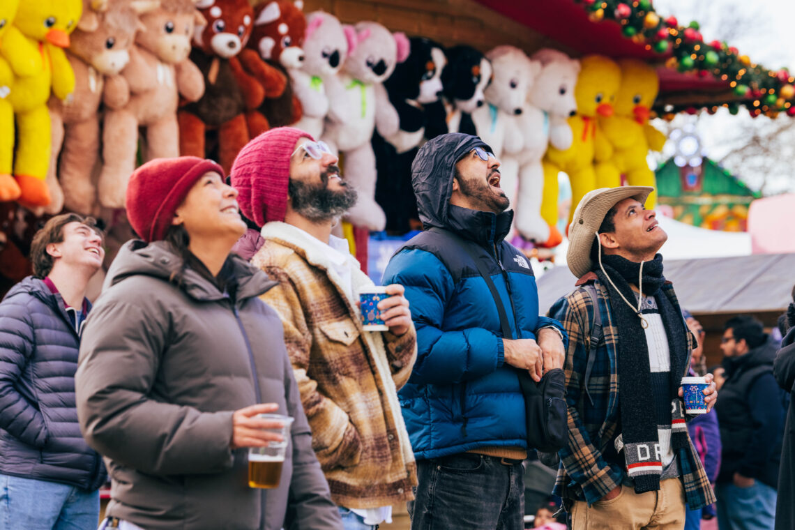 Group of men and women gazing at a ride in Winter Wonderland, holding drinks and wrapped up warm